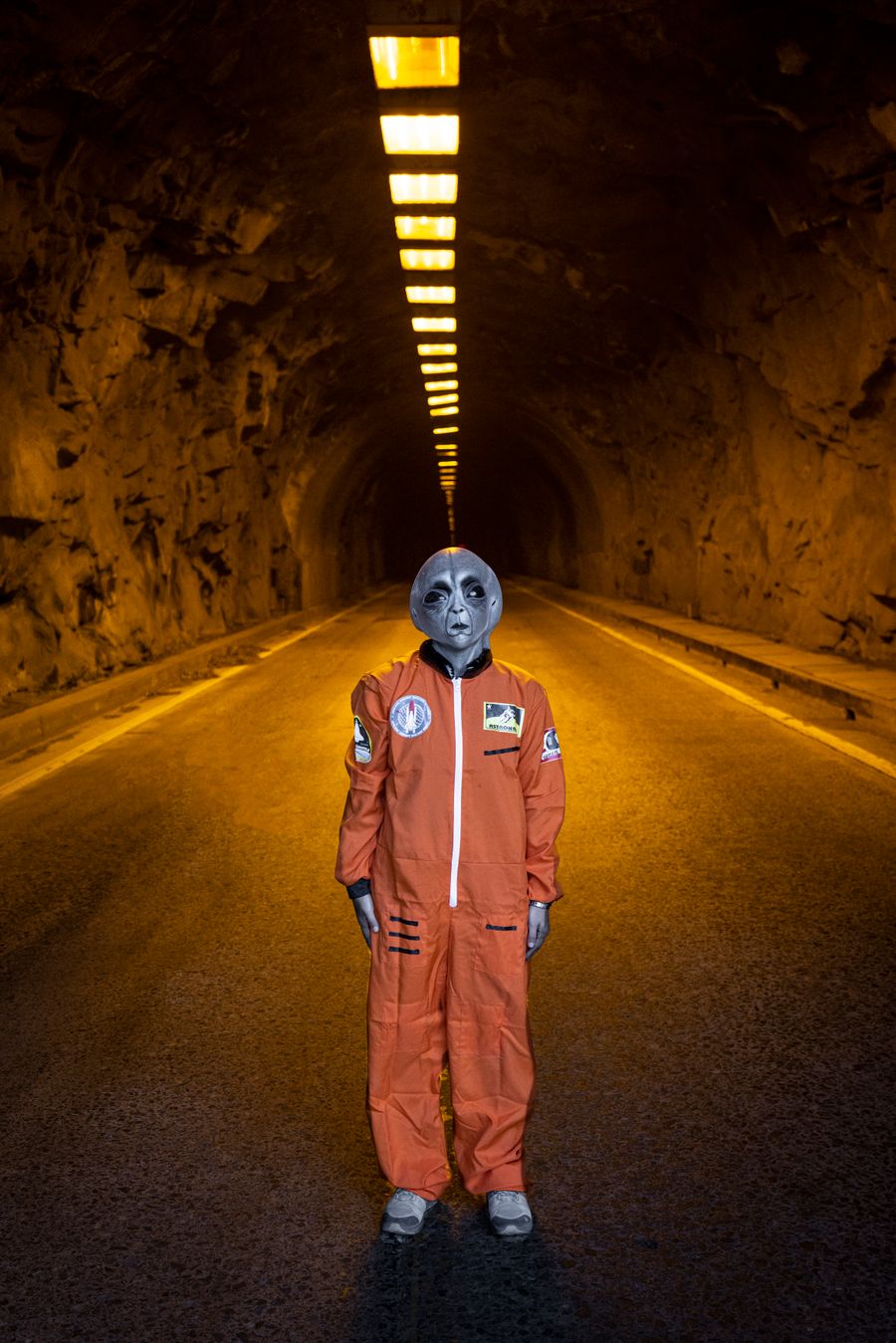 Visitante de máscara alienígena e traje espacial laranja, parado num túnel iluminado